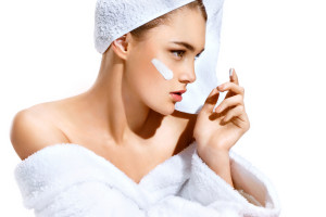 Young woman with flawless skin applying moisturizing cream on her face. Photo of woman after bath in white bathrobe and towel on white background. Skin care concept