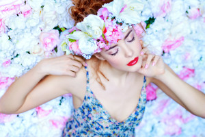 Beautiful romantic young woman in a wreath of flowers posing on a background of roses. Inspiration of spring and summer. Perfume, cosmetics concept.