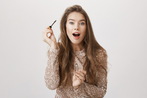 Emotive attractive caucasian woman looking at camera as if it is mirror with opened mouth while putting on mascara, standing in luxurious evening dress over gray background. Excited girl waits taxi.