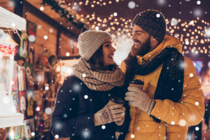 Having fun together at a christmas fairy with snowfall. Young cheerful couple is having a walk with hot drinks, enjoying, dressed warm, looking at each other and laugh, snowflakes all around