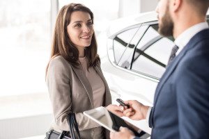 Portrait of beautiful young woman taking keys form car salesman standing next to white shiny luxury car in dealership showroom