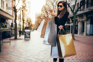 Shopper woman shopping with a smartphone in a city street. Caucasian female standing along the road holding shopping bags and using mobile phone.