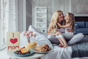 Attractive young woman with little cute girl are spending time together at home while sitting on bed. Happy family concept. Breakfast in bed on Mother's day.