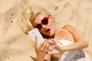 Technology and summer. Woman in red heart shaped sunglasses texting on mobile phone using smartphone reading sms or taking photo of herself on beach