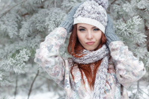 beautiful cheerful cute red-haired girl in a cap and scarf plays with snow in the beautiful forest fairy