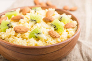 Millet porridge with kiwi and almonds in wooden bowl on a white wooden background and linen textile. Side view, close up, selective focus.