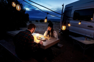 Young Couple Out Camping Deep In Concentration Playing Chess Together At A Picnic Table With Their Dog