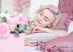 Young dreaming woman with closed eyes and short blond wavy bob haircut lying on pink bed linen at home. Square Pink Flower box with Fresh Rose is blurred in front.